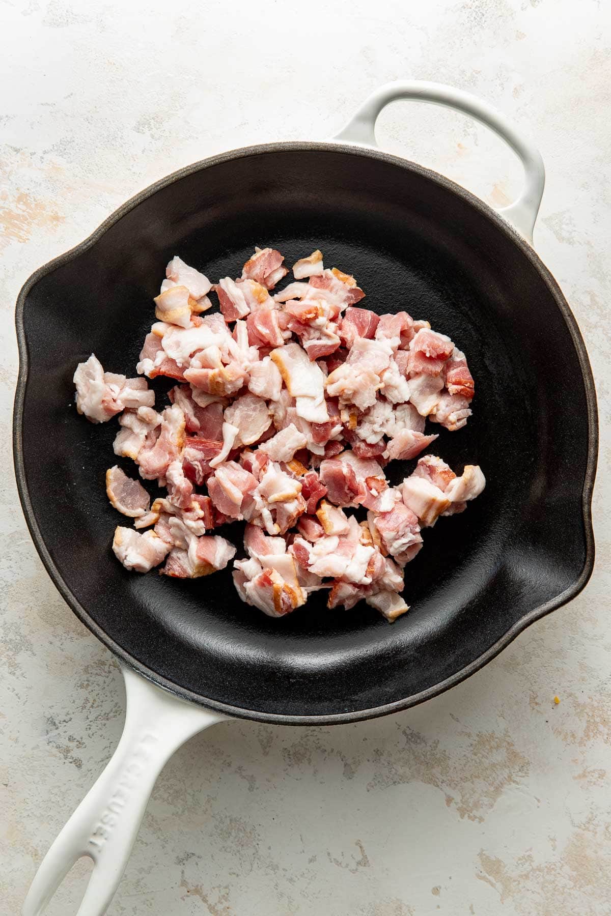 A white-handled cast iron skillet with chopped raw bacon pieces spread inside, placed on a light-colored countertop.