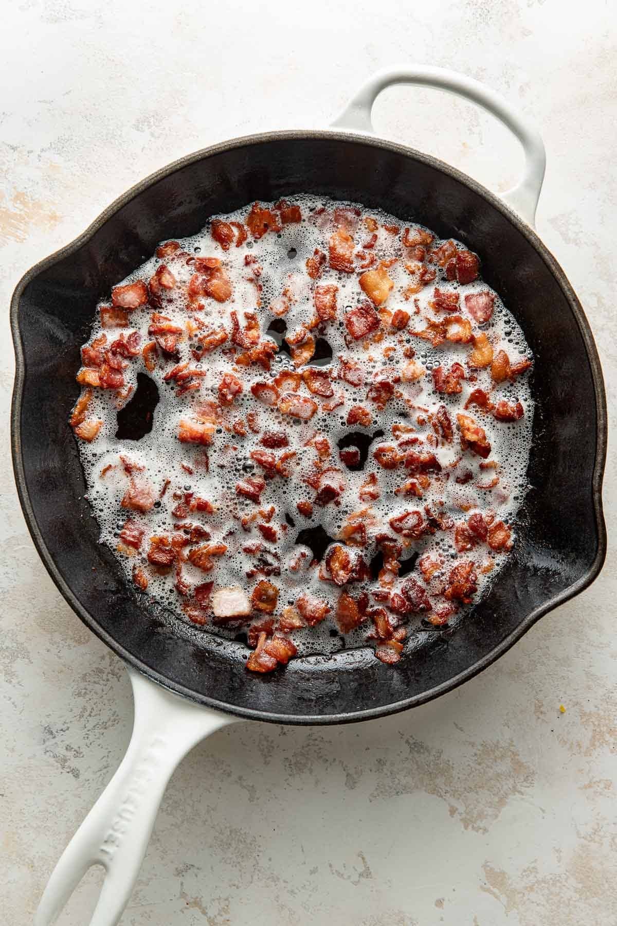 Crispy bacon pieces cooking in a foamy layer of fat inside a round black cast iron skillet with a white handle, viewed from above on a light countertop.