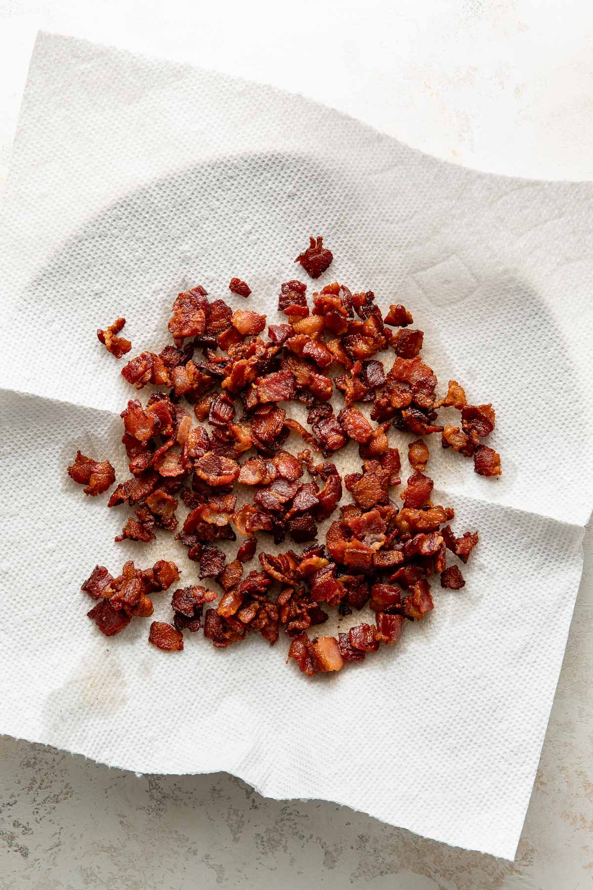 Chopped, crispy cooked bacon pieces sit on a white paper towel, which is placed on a white plate, absorbing excess grease.