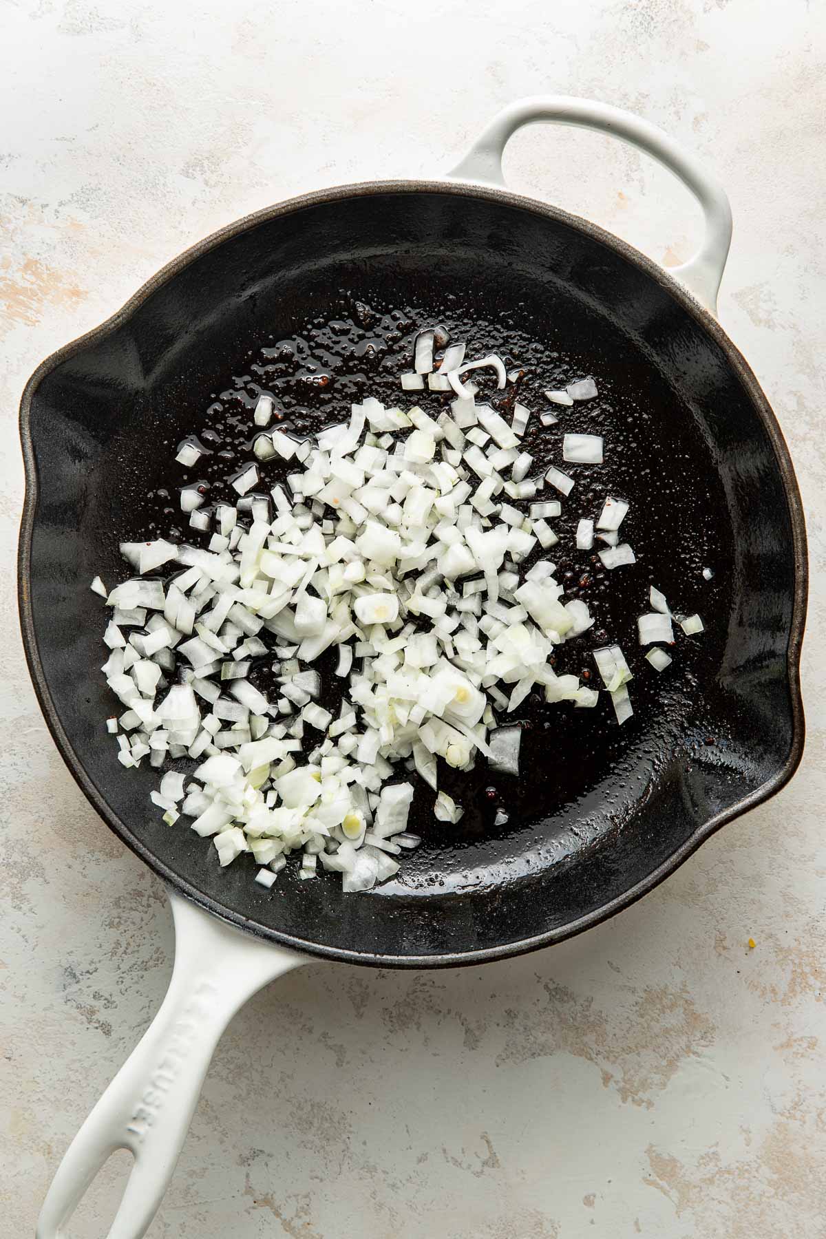 Chopped white onions in a white-handled black skillet on a light-colored countertop, ready to be cooked.