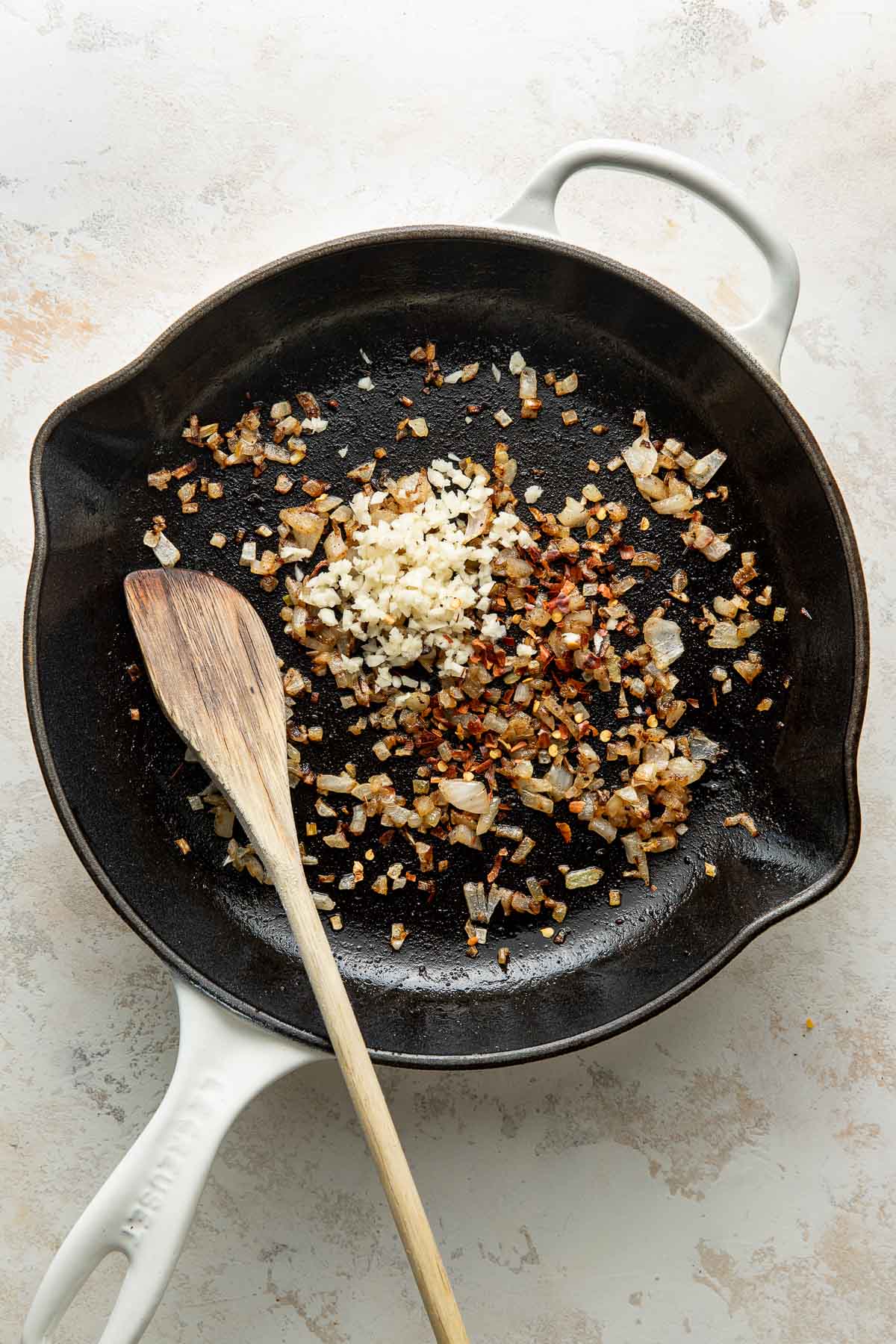 A white skillet with sautéed onions, garlic, and red pepper flakes, plus a wooden spoon resting inside.