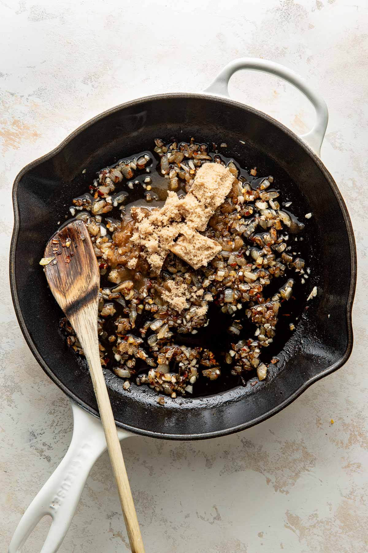 A white skillet with sautéed chopped onions, soy sauce, brown sugar, and a wooden spatula resting inside, seen from above on a light surface.