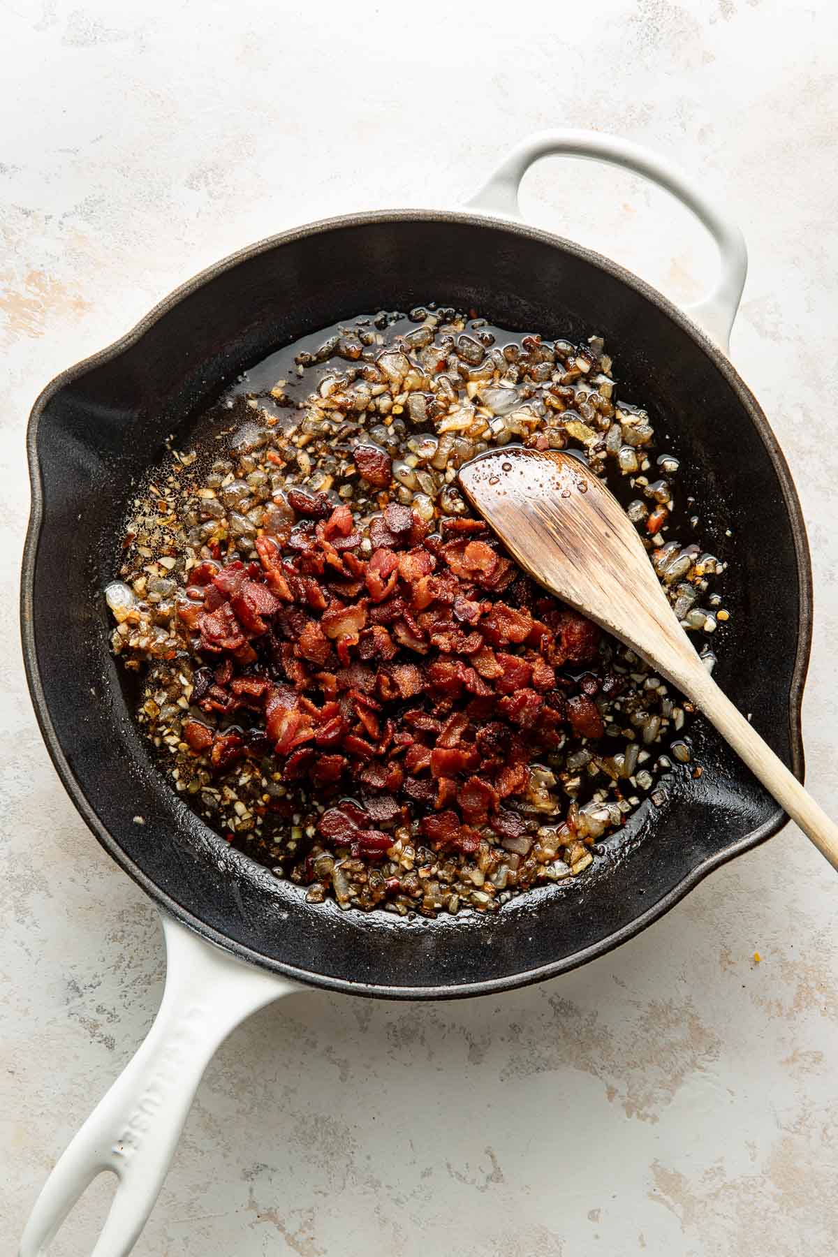 A black skillet with sautéed chopped bacon, onions, and garlic, being stirred with a wooden spoon on a light countertop.