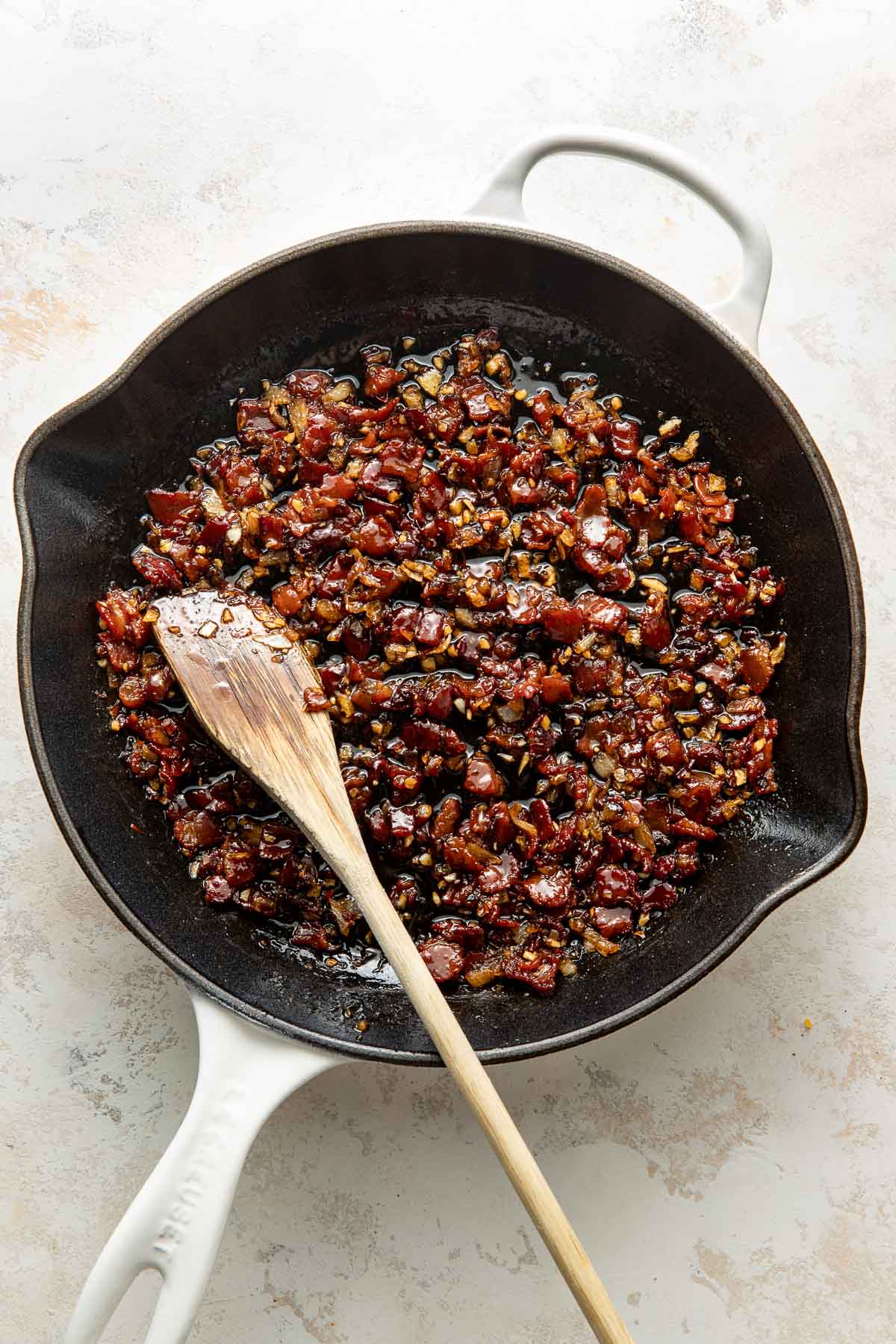 A cast iron skillet filled with cooked, diced bacon and onions, with a wooden spoon resting in the mixture. The skillet sits on a light-colored surface.