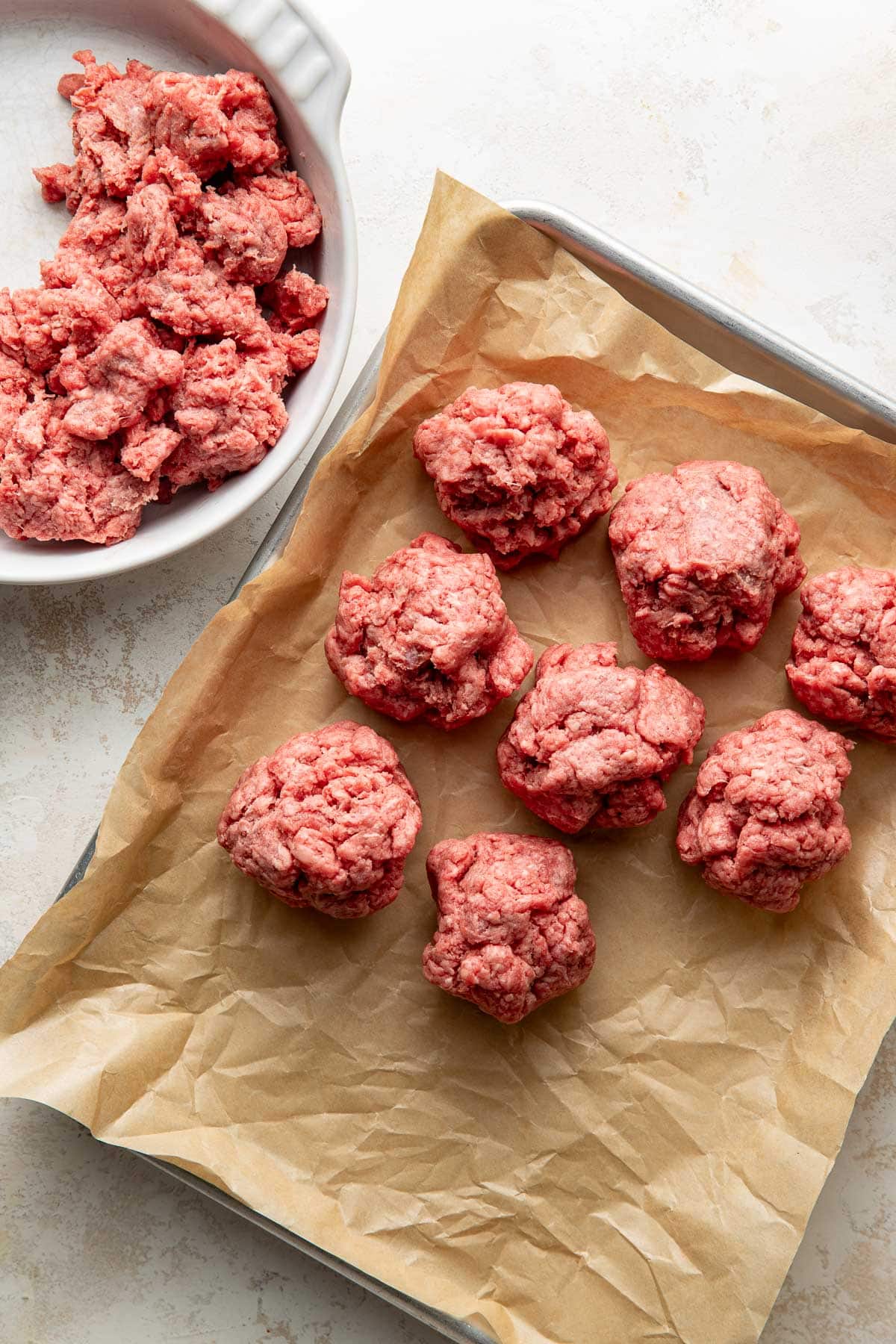 Raw ground beef shaped into balls sits on a baking sheet lined with parchment paper, with a bowl of loose ground beef nearby on a light surface.