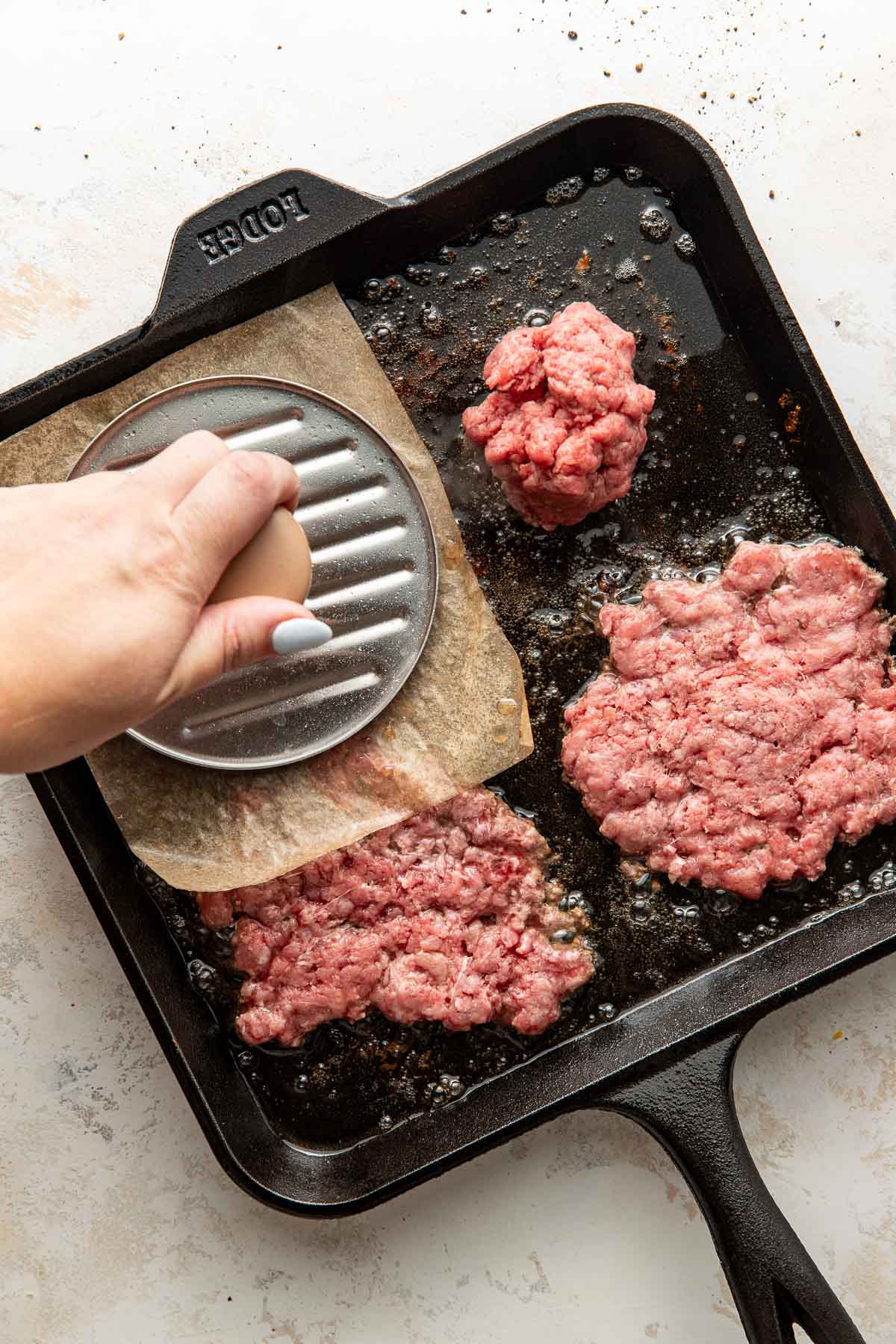 A hand uses a metal press with parchment paper to flatten ground beef on a hot cast iron skillet, forming burger patties.