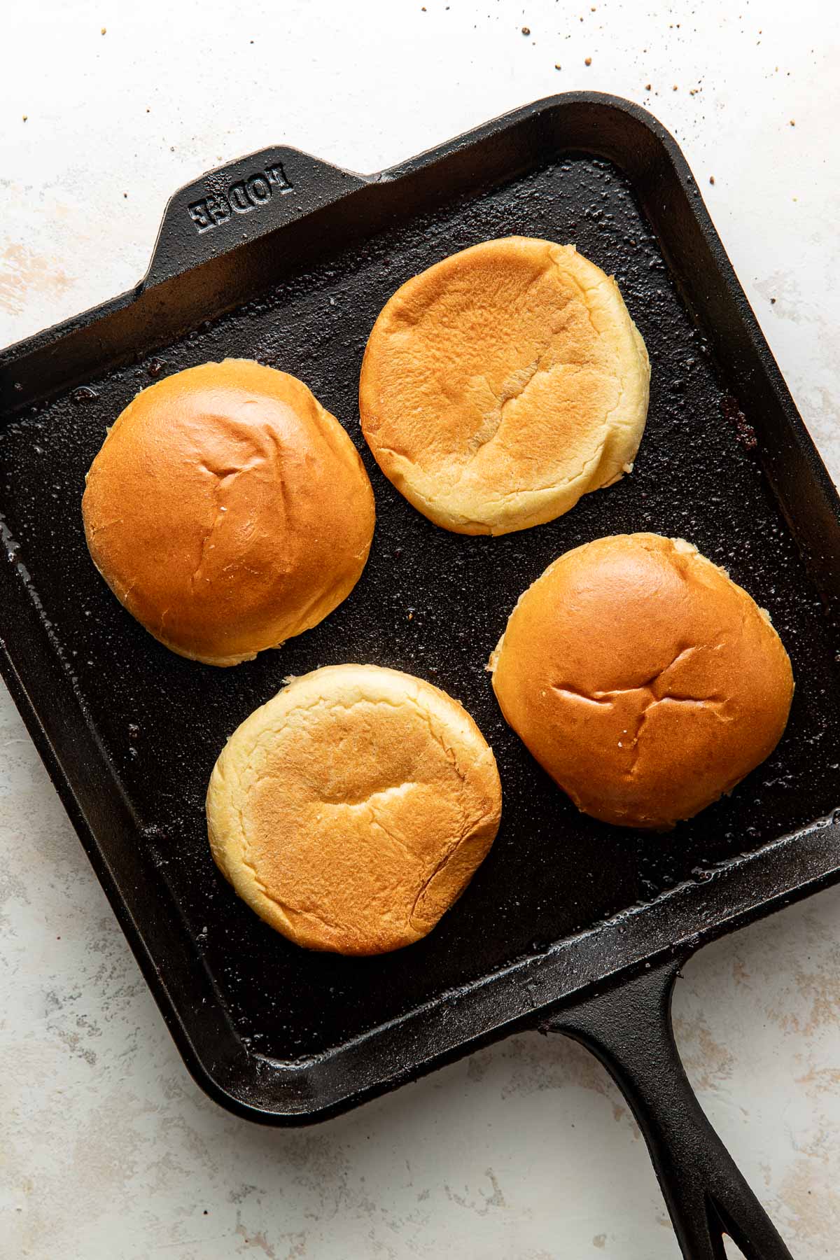 Four hamburger buns, cut side down, toasting on a black cast iron skillet. The buns are golden brown and placed on a white countertop.