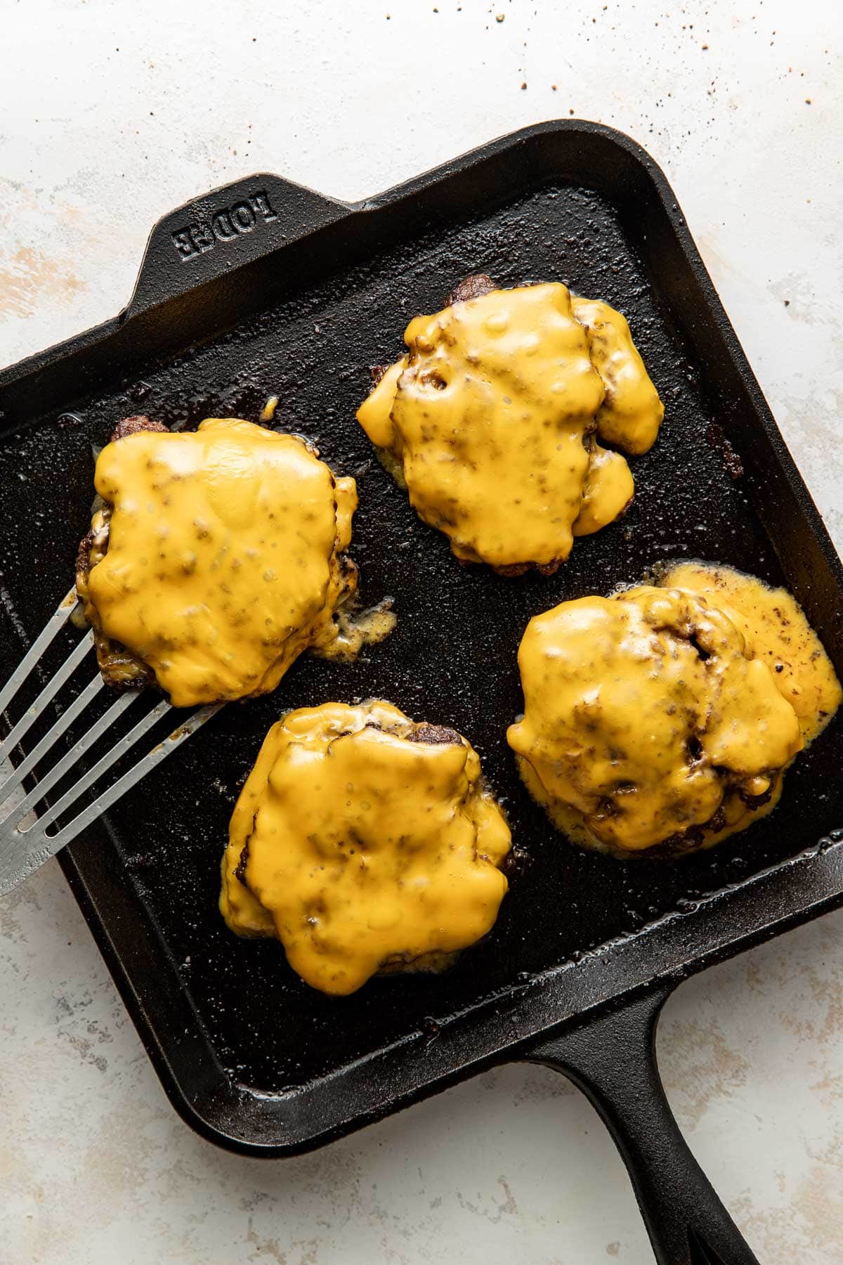 Four cheeseburgers with melted cheese on top are cooking on a black cast iron grill pan. One burger is being lifted with a metal spatula. The background is a light-colored surface.