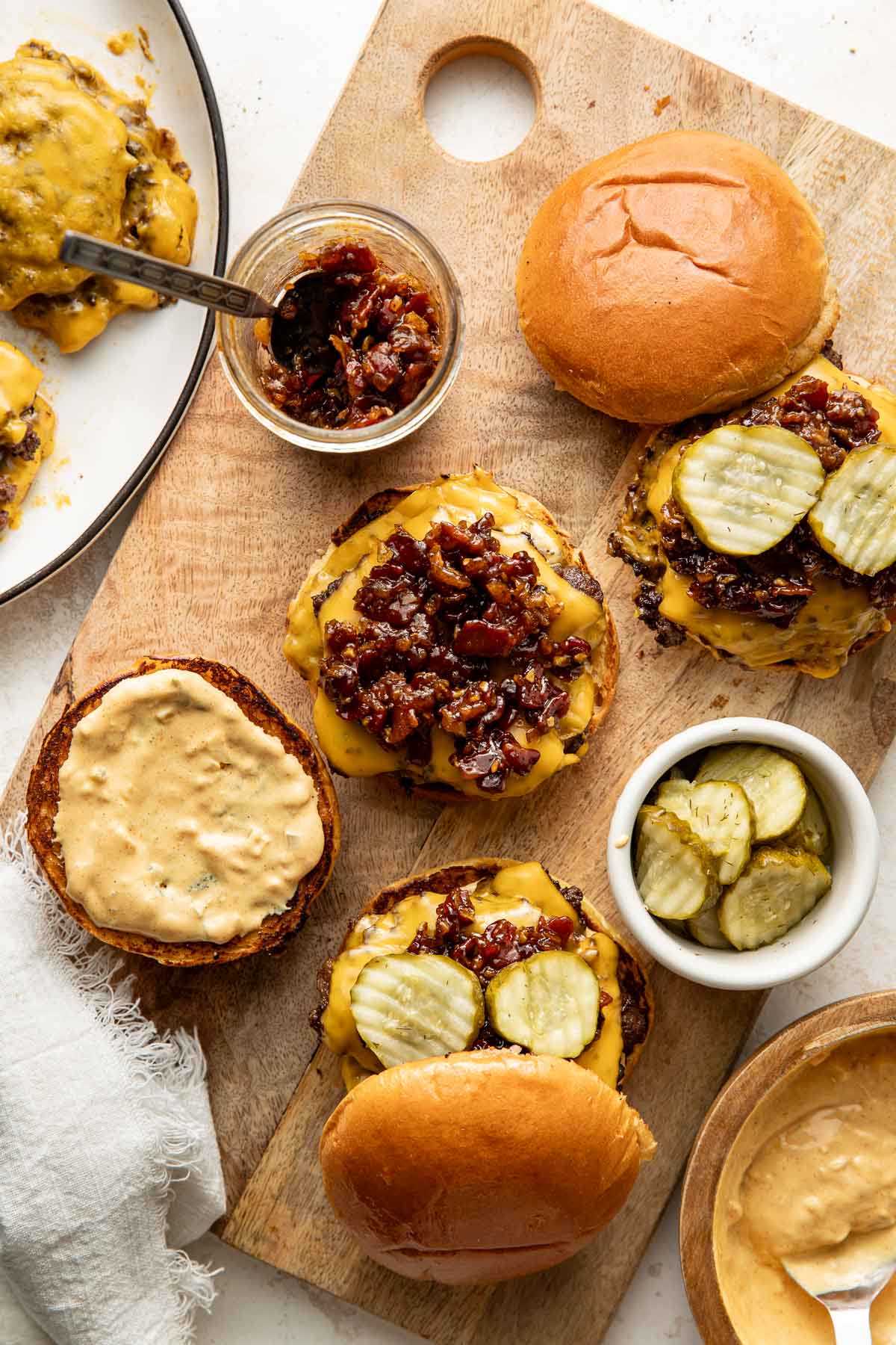 Cheeseburgers on a wooden board topped with pickles and relish, surrounded by buns, a jar of relish, a bowl of pickles, and a plate of more burgers.