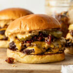 A cheeseburger with melted cheese, pickles, crispy bacon bits, and sauce on a toasted bun, resting on a wooden surface. Another similar burger is blurred in the background.