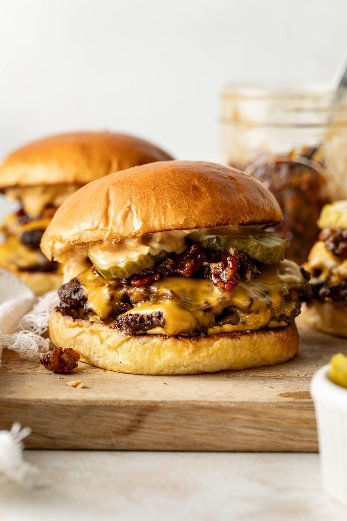 A cheeseburger with melted cheese, pickles, bacon, and sauce in a brioche bun sits on a wooden board. Two more burgers and a jar of sauce are blurred in the background.