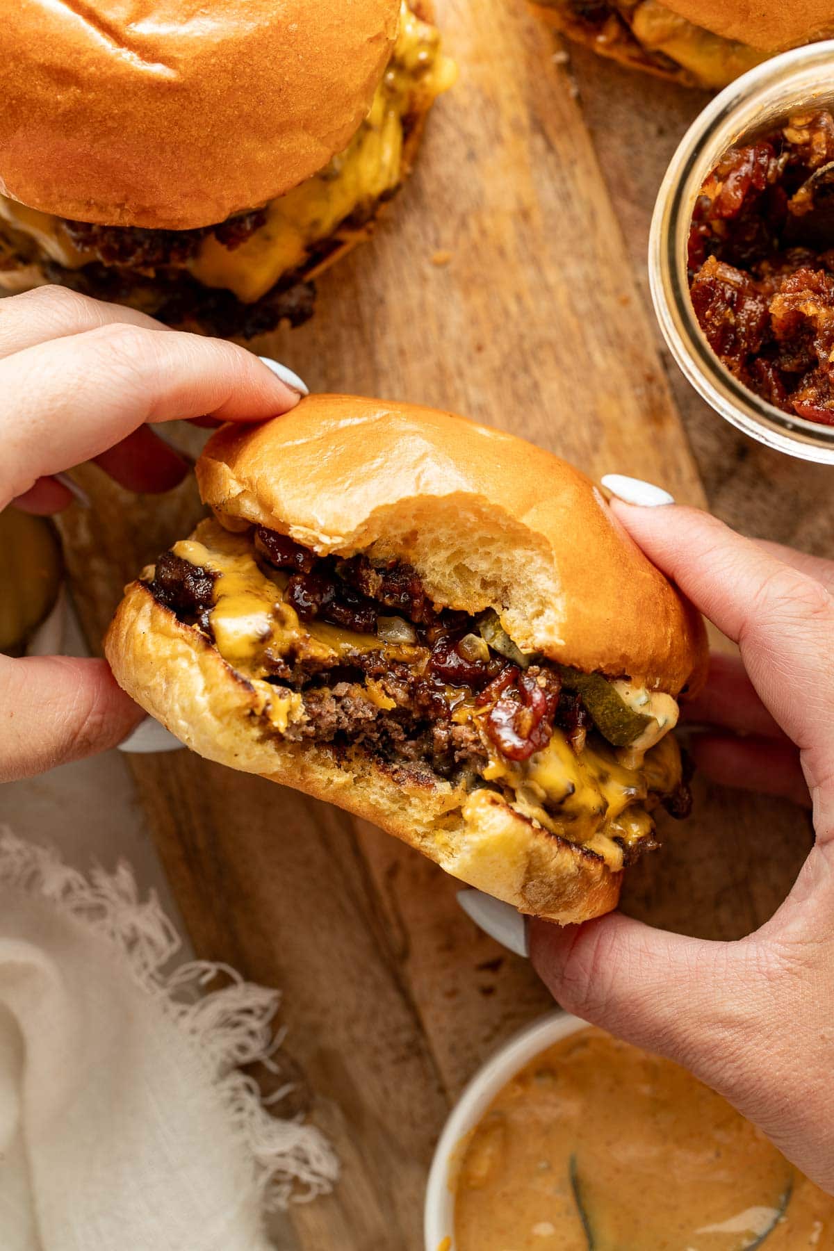 A person holding a cheeseburger with a bite taken out, showing melted cheese, pickles, and what appears to be bacon jam, on a wooden surface with condiments and another burger nearby.