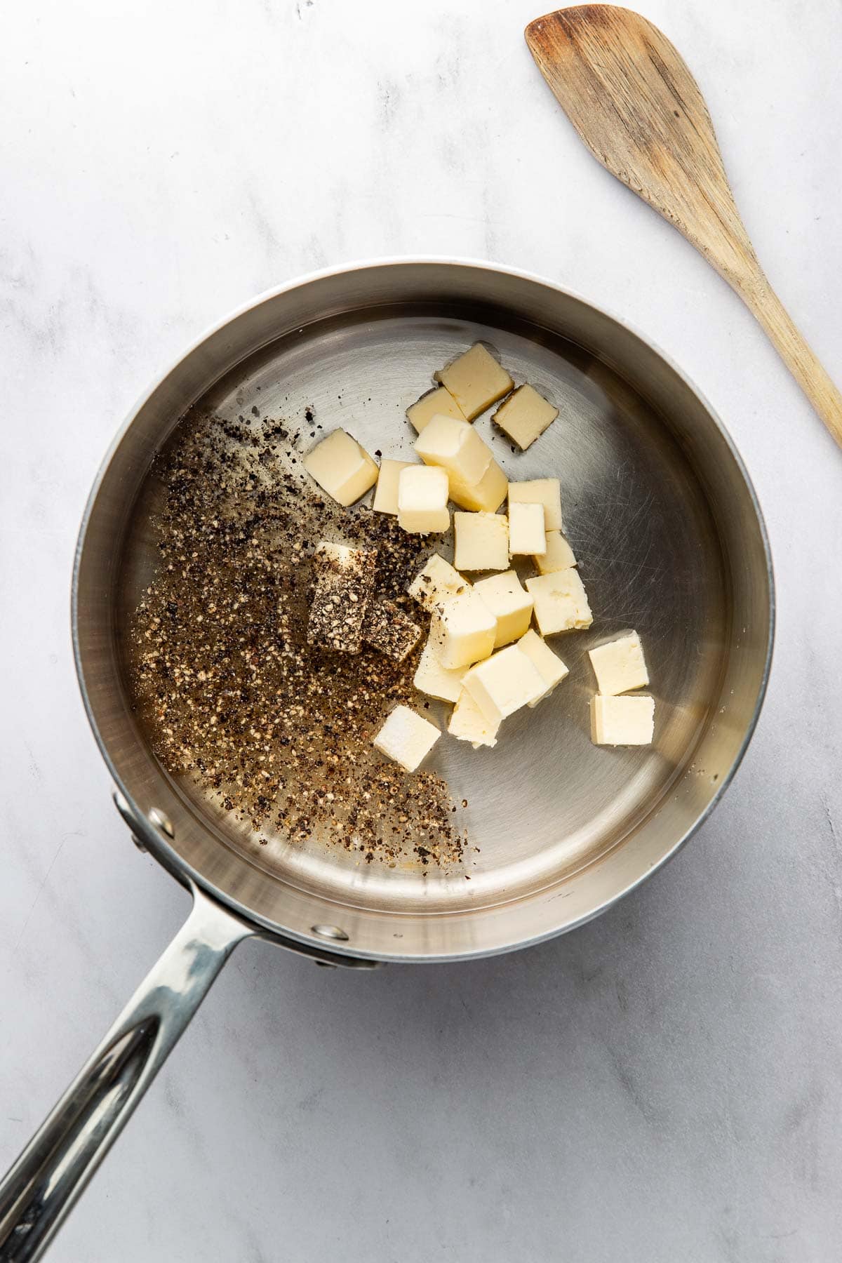 A stainless steel saucepan with cubed butter and cracked black pepper inside, next to a wooden spoon on a white surface.