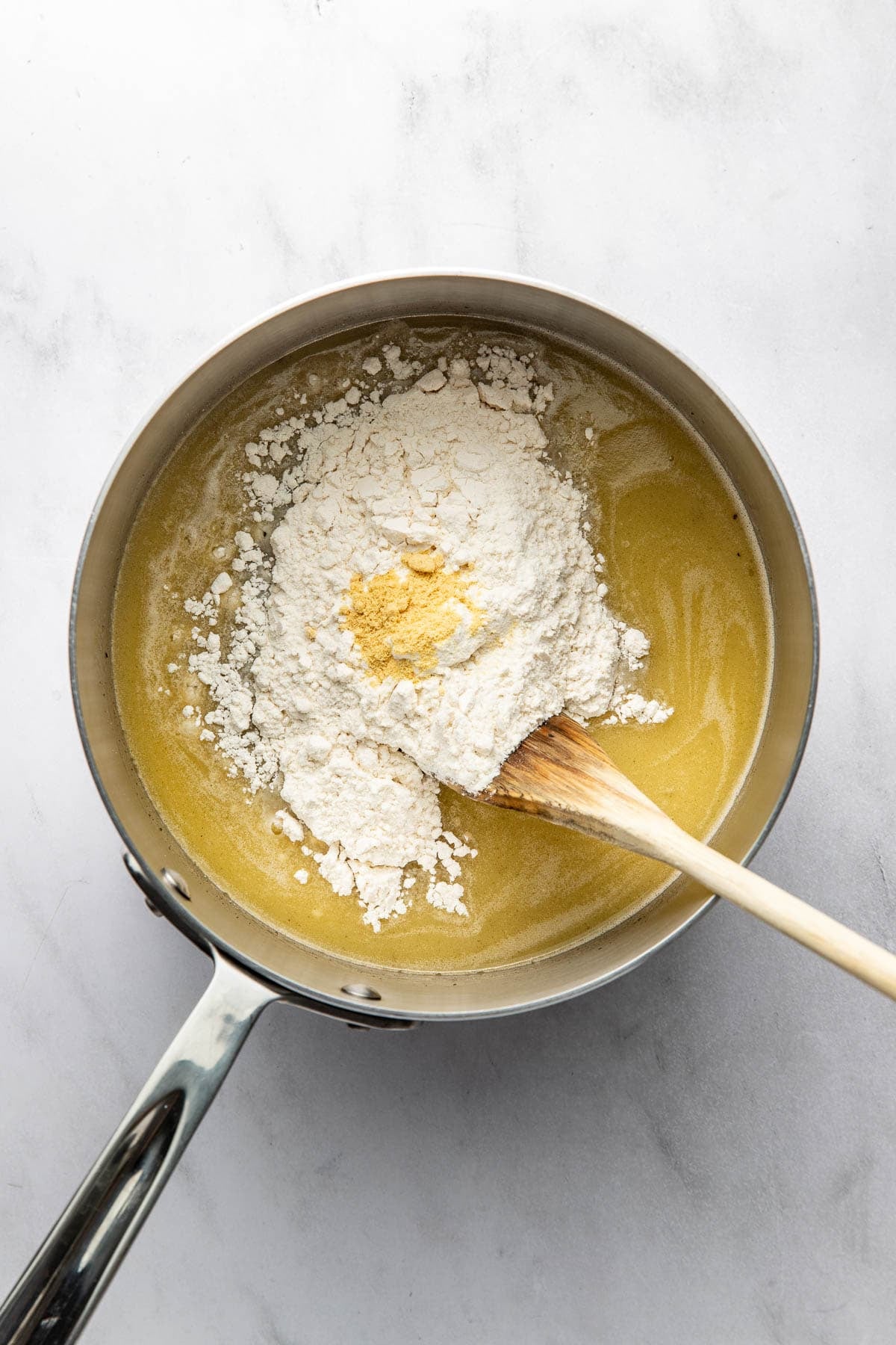 A saucepan with melted butter, flour, and a bit of yellow seasoning, being stirred with a wooden spoon on a white marble surface.