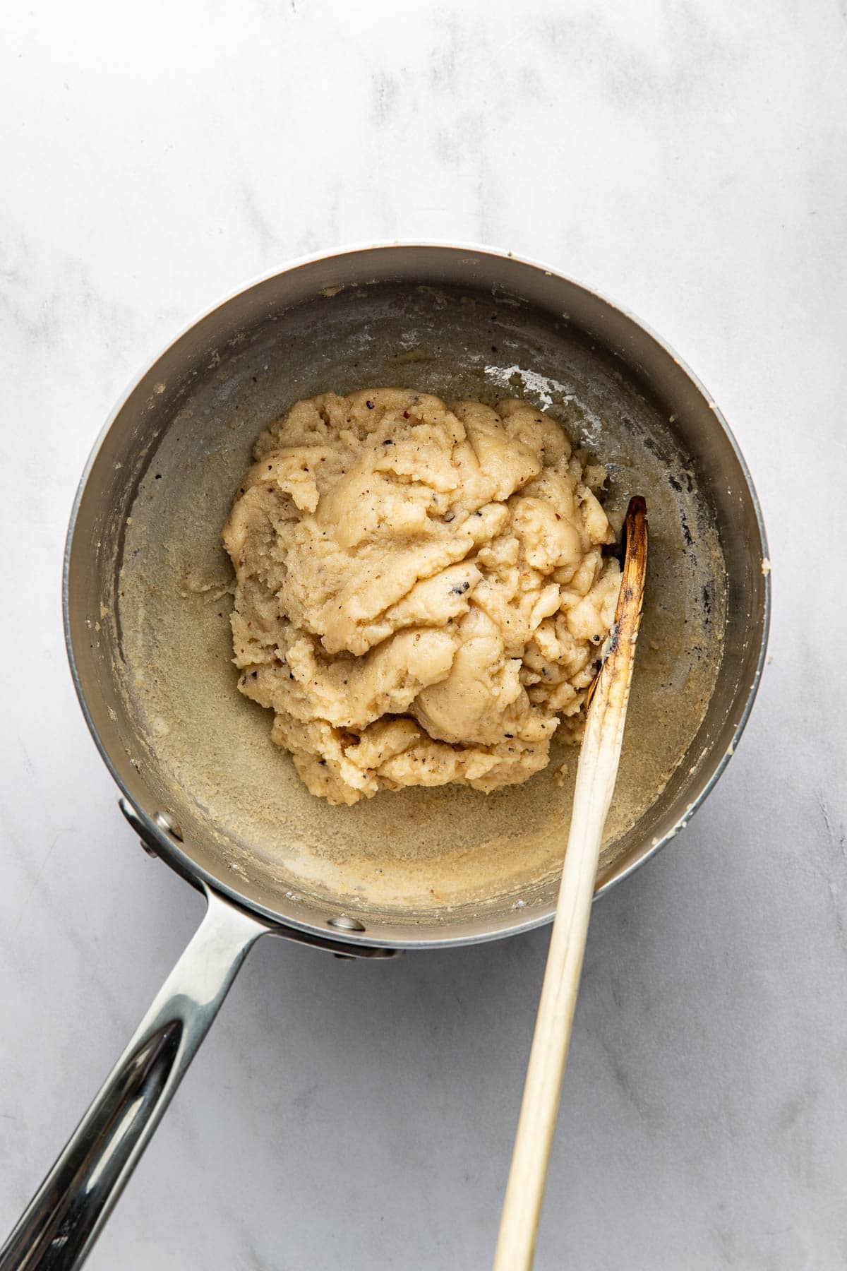 A saucepan with a thick, beige dough mixture inside and a wooden spoon resting on the edge, all set on a white marble surface.