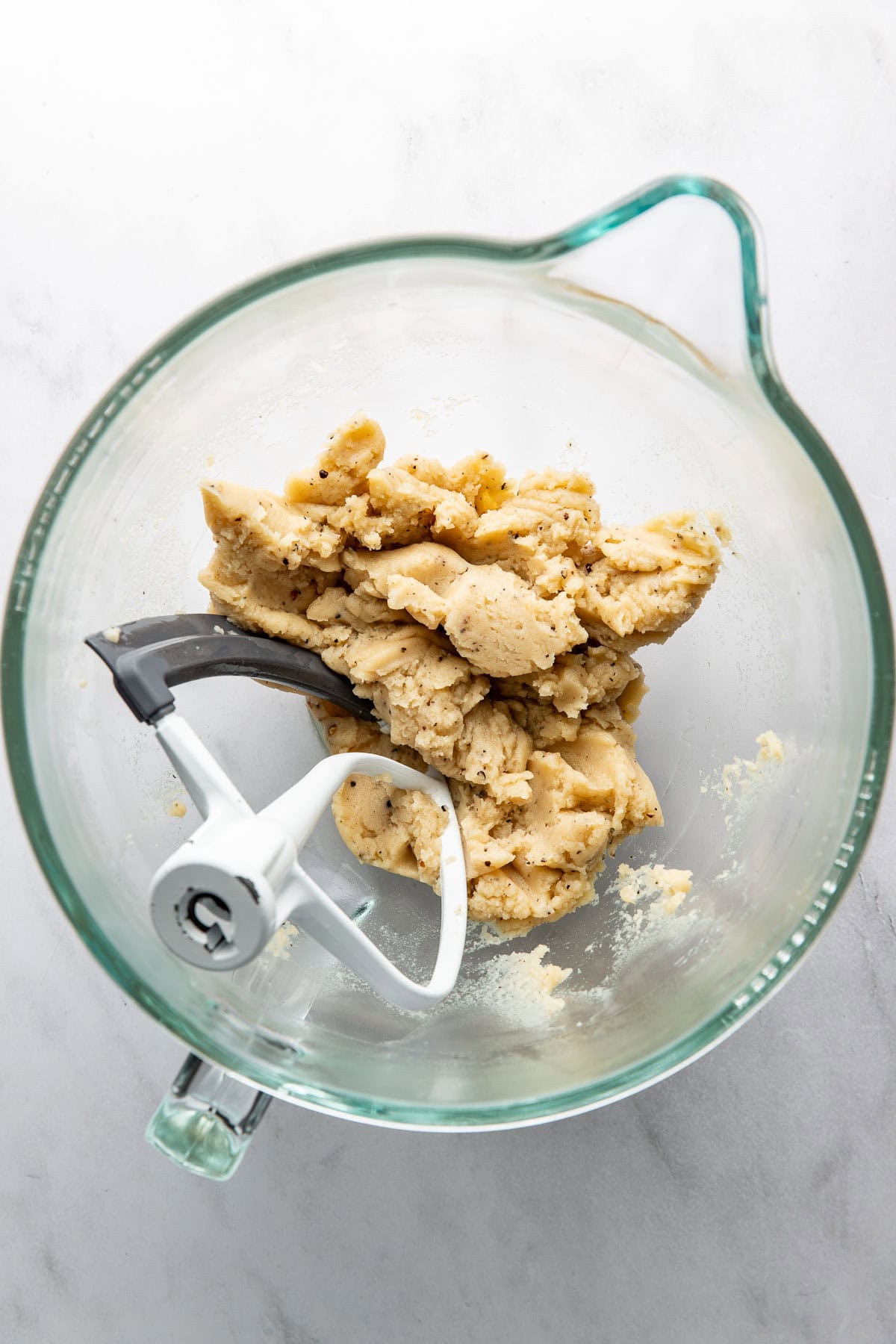 A glass mixing bowl with a metal and plastic paddle attachment contains a lump of pale cookie dough on a light marble surface.