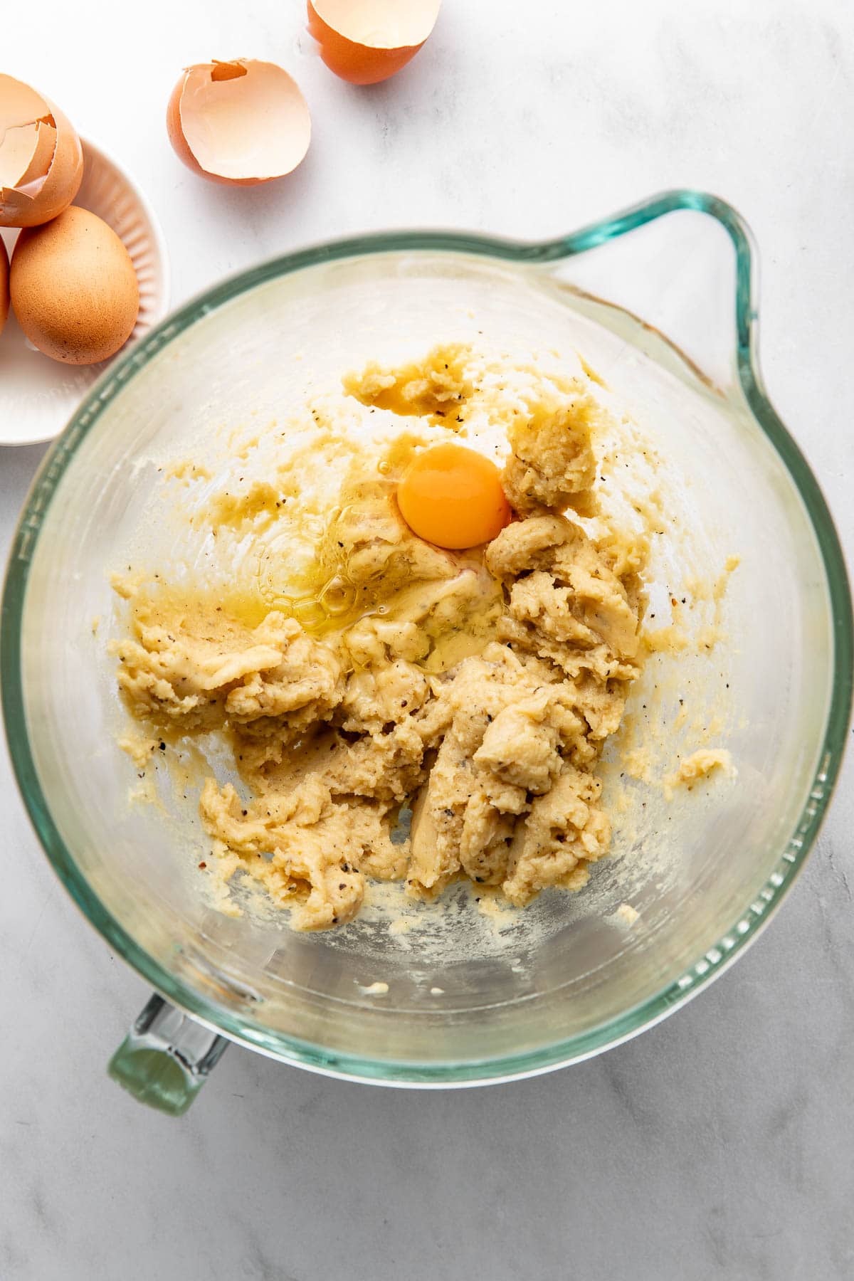 A glass mixing bowl with cookie dough and a cracked egg inside. Eggshells and whole eggs are on a small plate nearby, all on a white countertop.