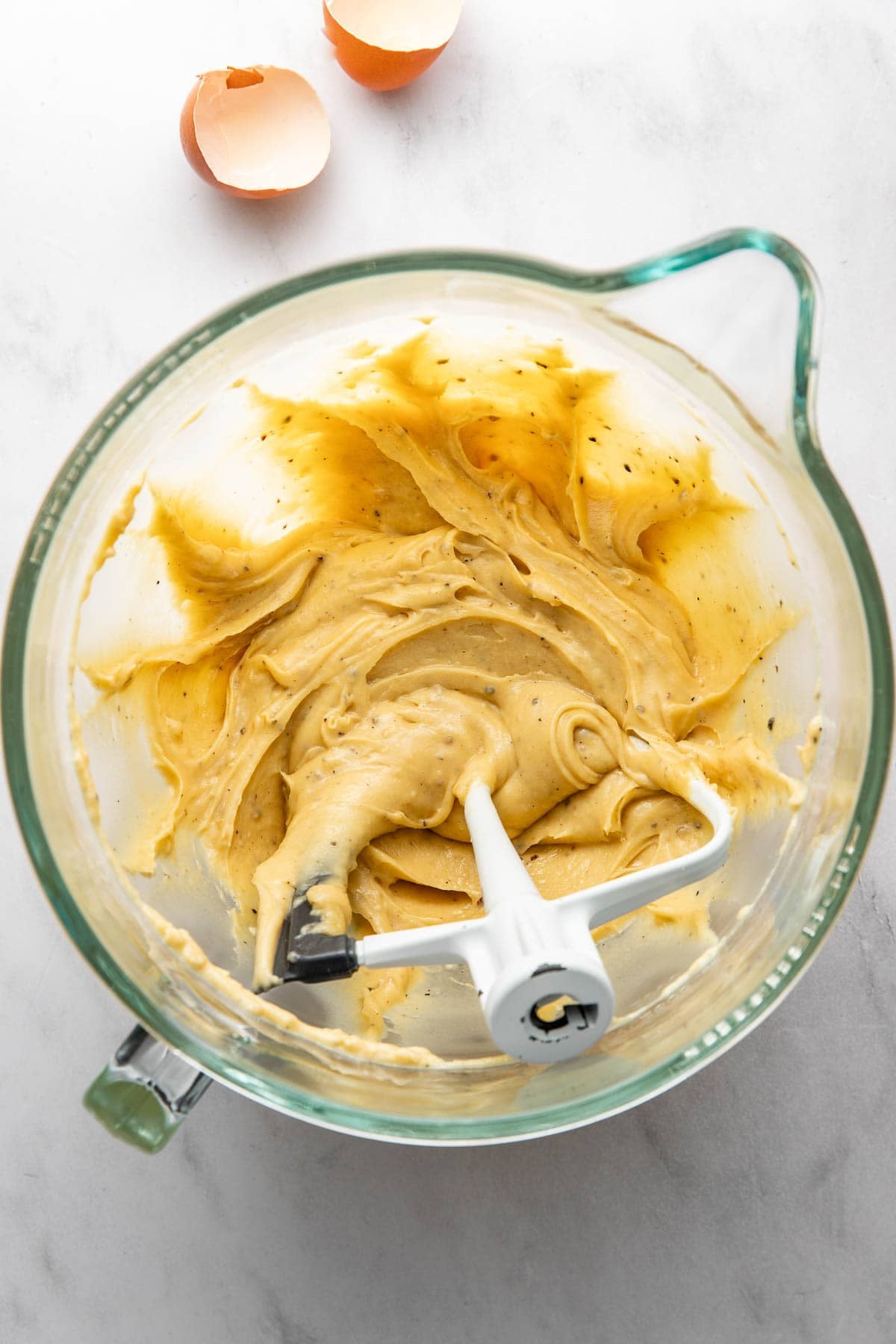 A glass mixing bowl with cookie dough and a mixer paddle inside, seen from above. Two cracked egg shells rest beside the bowl on a light-colored surface.