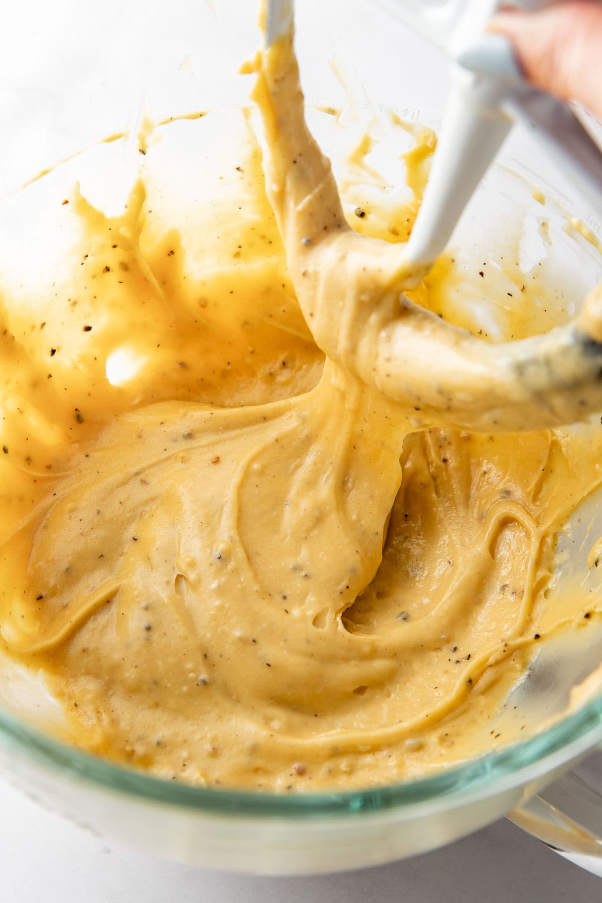 A glass mixing bowl filled with thick, yellow batter speckled with black pepper, with a stand mixer paddle attachment lifting some of the batter.