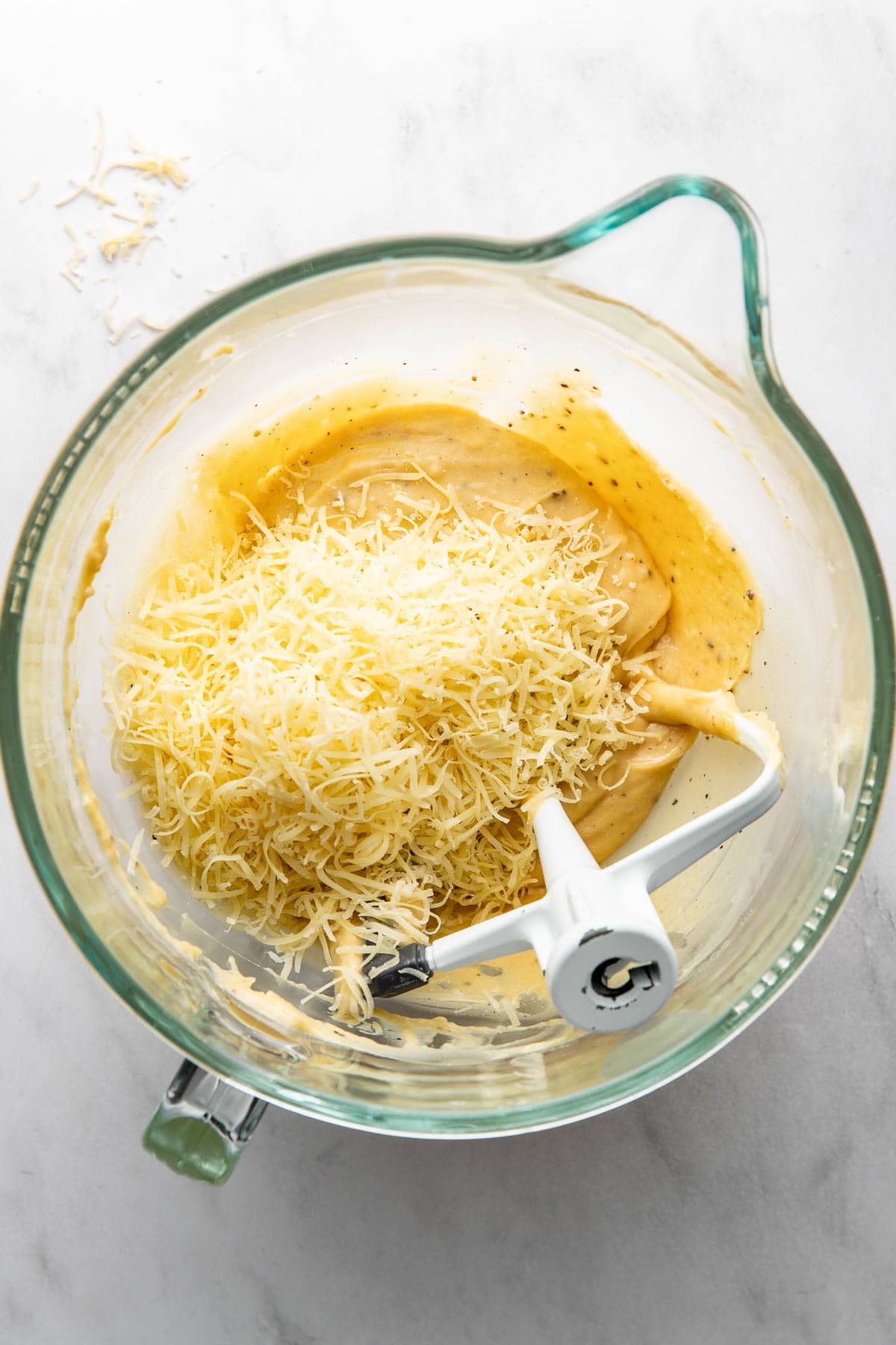 A glass mixing bowl with shredded cheese and batter, partially mixed with a stand mixer paddle attachment on a white countertop.