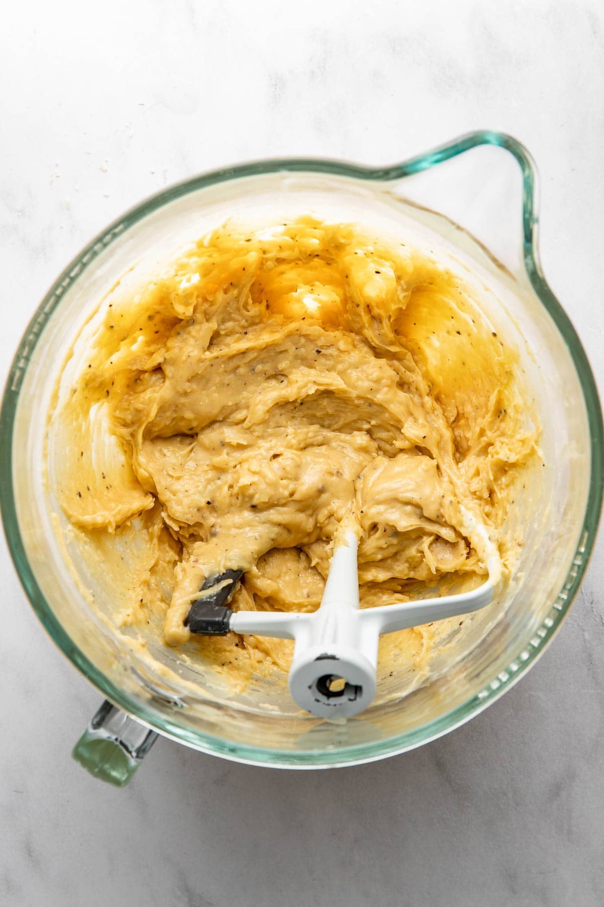 A glass mixing bowl on a white surface contains creamy cookie dough and a paddle attachment, with some dough clinging to the sides.