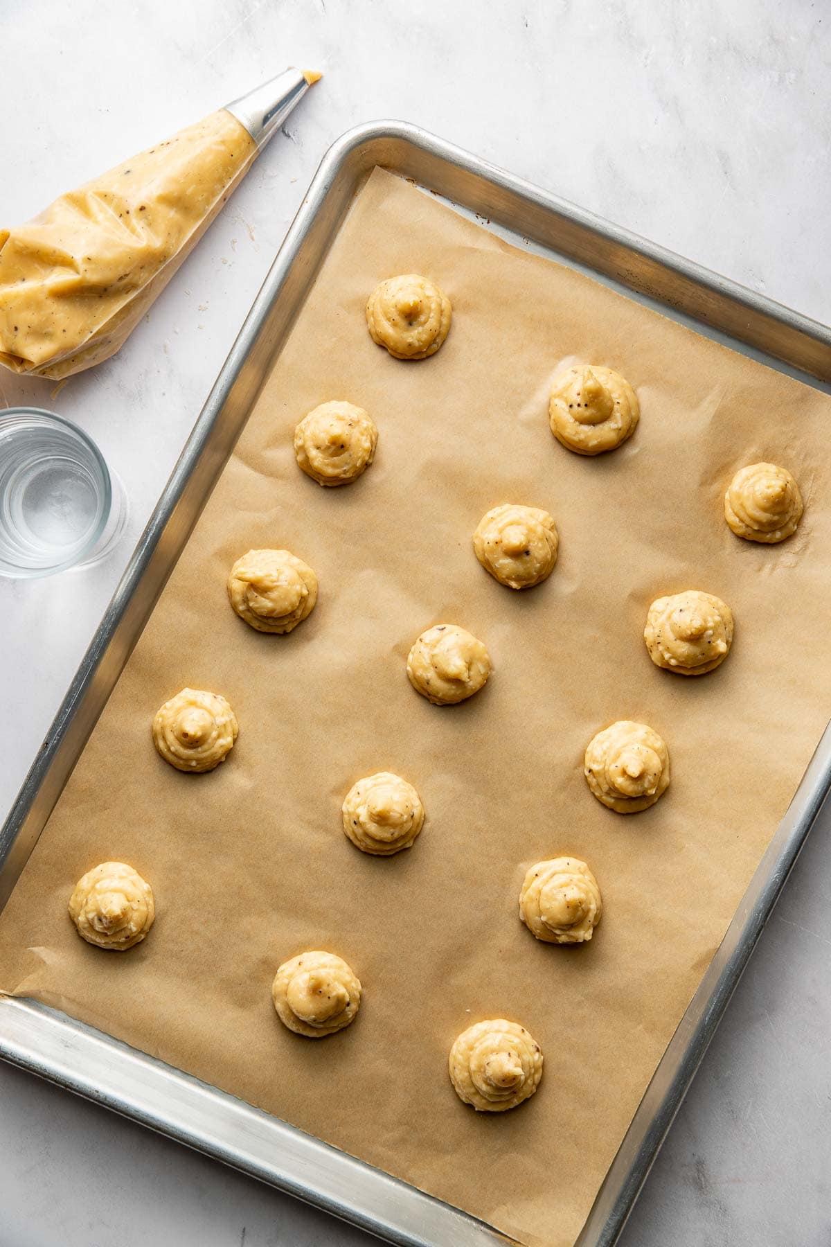 A baking tray lined with parchment paper holds rows of piped dough. A piping bag and a glass of water are placed nearby on a white surface.
