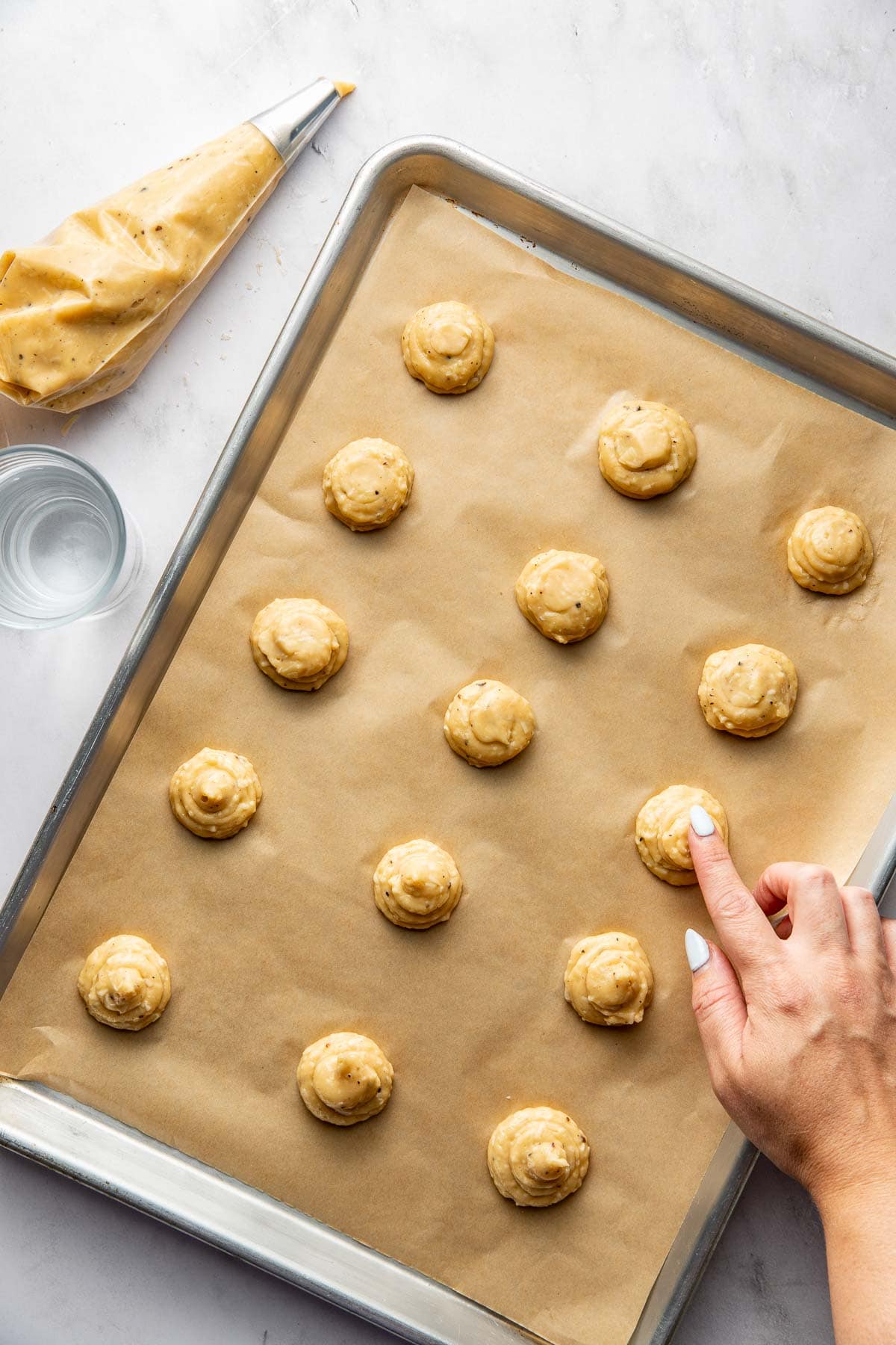 A hand shapes dough mounds on a parchment-lined baking sheet. A piping bag and a glass of water are nearby on a white surface.