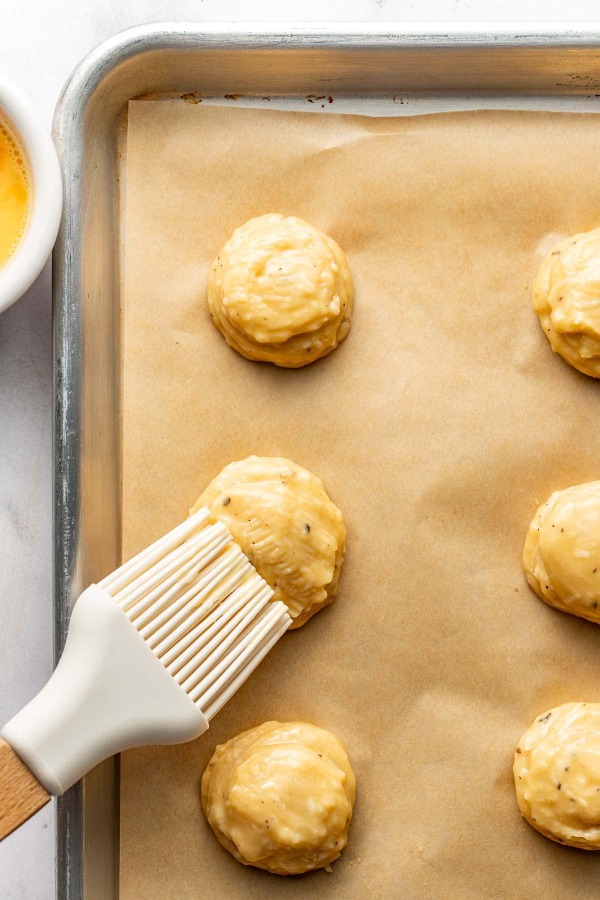 A pastry brush applies egg wash to round dough balls on a parchment-lined baking sheet. A bowl of egg wash is partially visible in the corner.