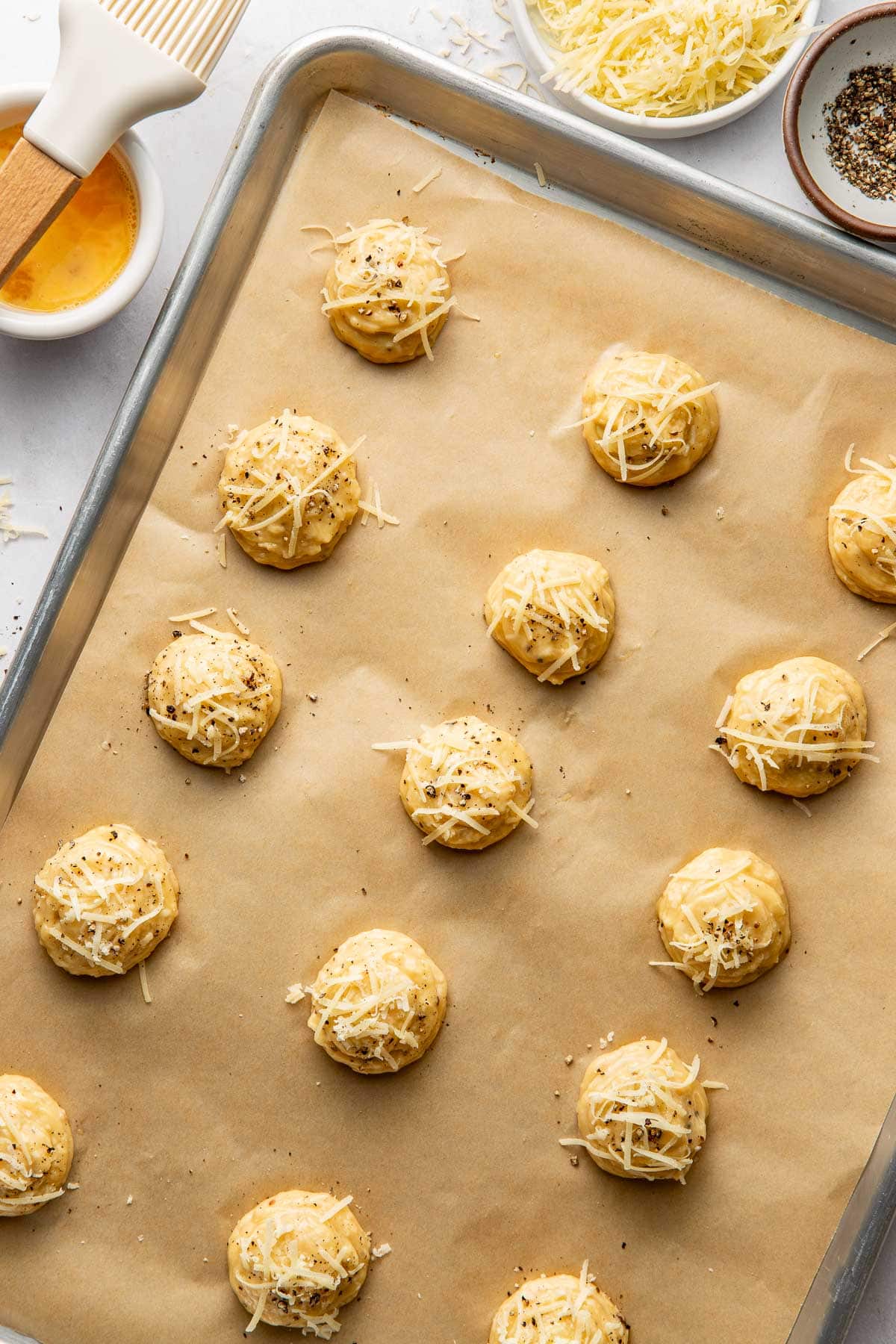 Unbaked cheese-topped dough balls arranged on a parchment-lined baking sheet, with bowls of grated cheese, egg wash, a basting brush, and pepper nearby.