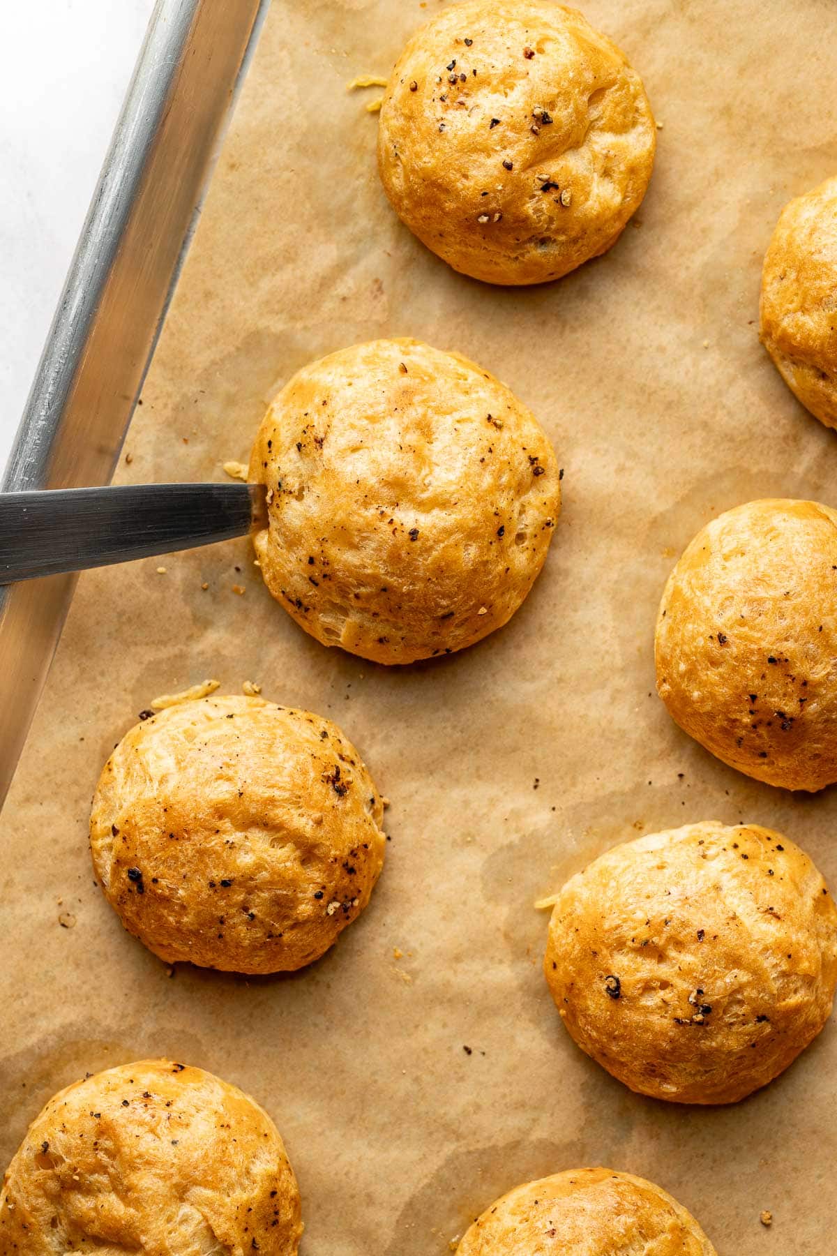 Round, golden-brown cheese puffs on parchment paper, sprinkled with black pepper. A metal spatula is sliding under one puff, ready to lift it from the baking tray.