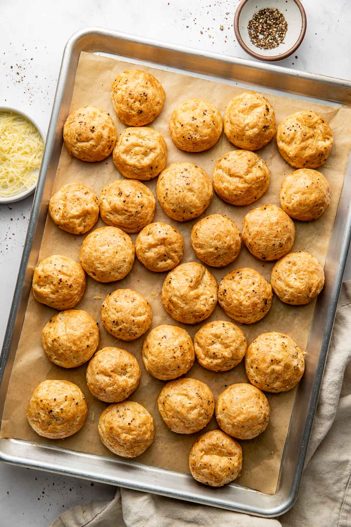 A baking sheet lined with parchment paper holds 28 golden-brown cheese puffs. Nearby are a bowl of shredded cheese, a small bowl of pepper, and a beige cloth.