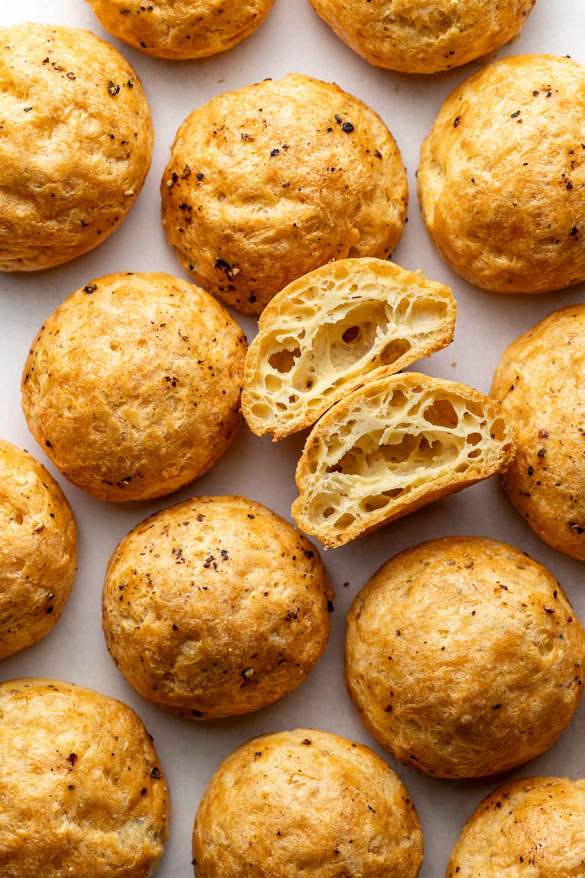 Round, golden-brown cheese puffs are arranged on a white surface. One puff is cut in half, revealing a light, airy, and hollow interior. Black pepper flecks are visible on the tops of the puffs.