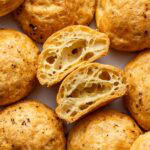 Round, golden-brown cheese puffs with a crispy exterior. One puff is cut in half, showing a hollow, airy interior. Black pepper flecks are visible on the surface.