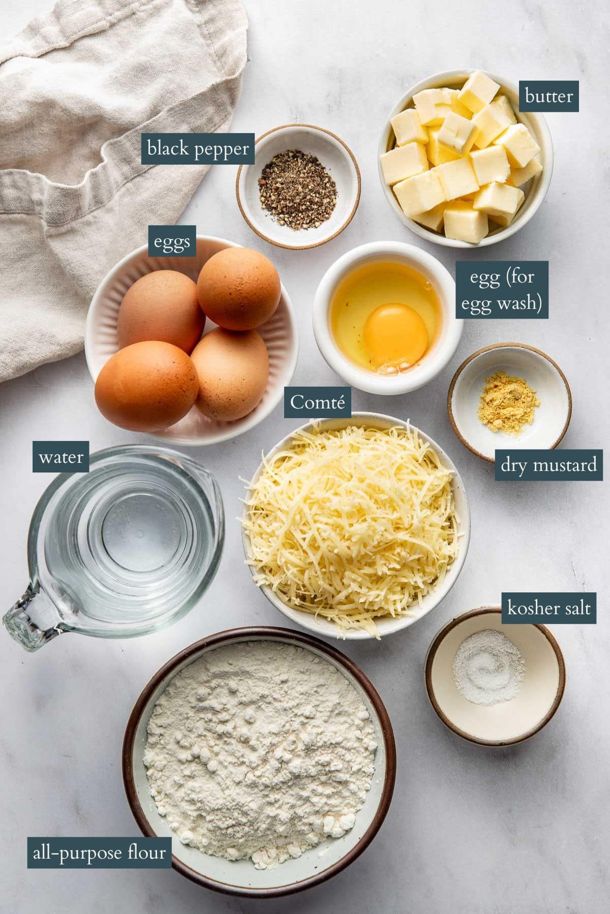 Various labeled ingredients for baking are arranged on a white surface, including eggs, butter, shredded cheese, flour, water, kosher salt, black pepper, dry mustard, and a cracked egg for egg wash.