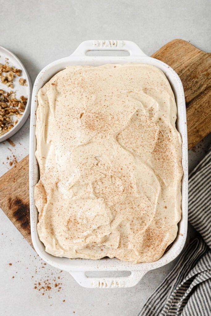 A white ceramic dish filled with a creamy dessert topped with cinnamon, resting on a wooden board beside a small plate of chopped nuts and a striped cloth.