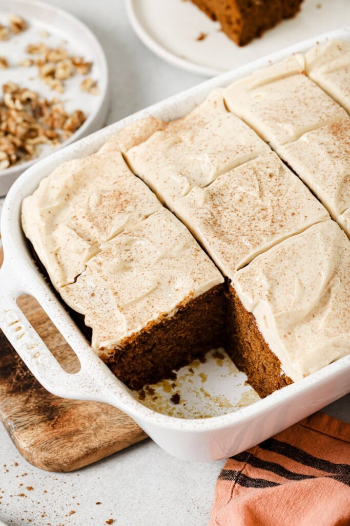A white baking dish with frosted cake cut into squares, one piece removed. Nearby are a bowl of chopped nuts, a wooden board, and an orange-striped cloth.