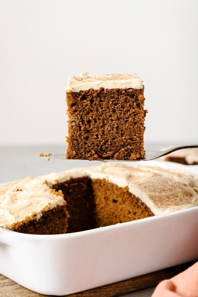 A fork lifts a square slice of frosted gingerbread cake from a white baking dish, showing the cakes moist texture and light brown frosting on top.