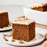 A square slice of spiced cake with frosting and walnut pieces on top sits on a white plate, with a fork and more cake pieces in the background.