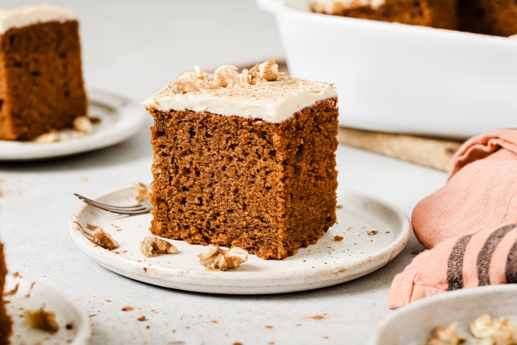 A slice of spiced cake with creamy frosting and walnuts on top sits on a white plate, with a fork and scattered walnut pieces nearby. Other slices and a baking dish are in the background.