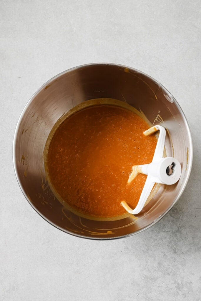 A metal mixing bowl containing an orange batter with a paddle attachment resting inside, set on a light gray surface.