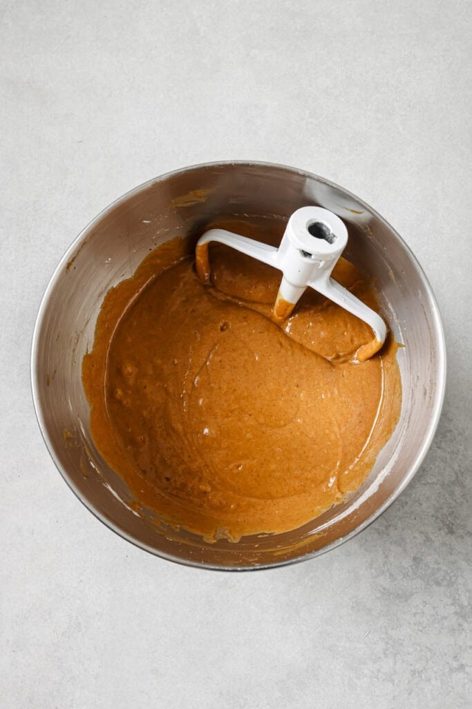 A mixing bowl filled with light brown batter and a white paddle attachment, resting on a grey countertop.