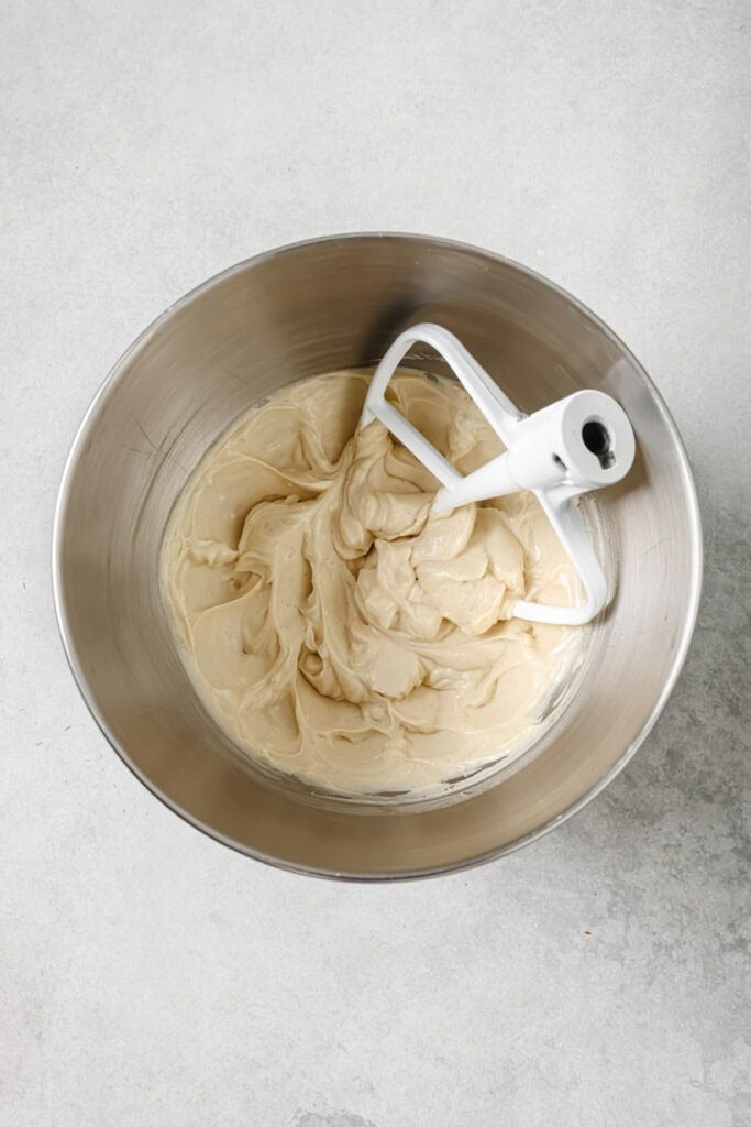 A metal mixing bowl with light beige cake batter and a white paddle attachment resting inside, viewed from above on a gray surface.