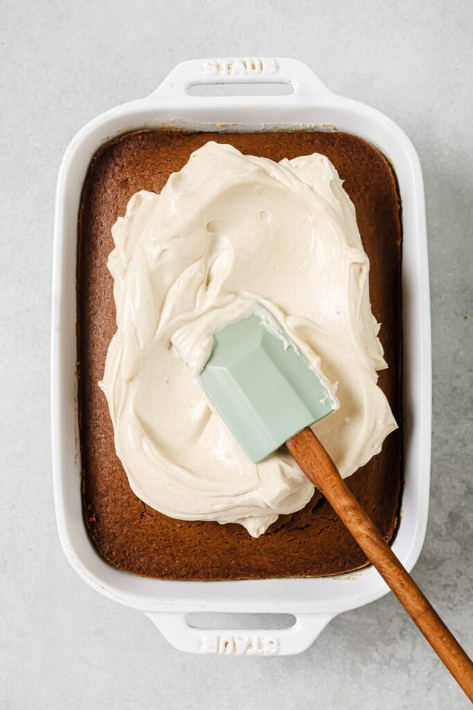 A baked sheet cake in a white dish with thick white frosting being spread on top using a mint green spatula with a wooden handle.