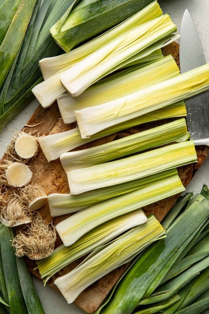 Several cleaned and cut leeks are arranged on a wooden cutting board with a knife, leek roots, and outer leaves nearby.