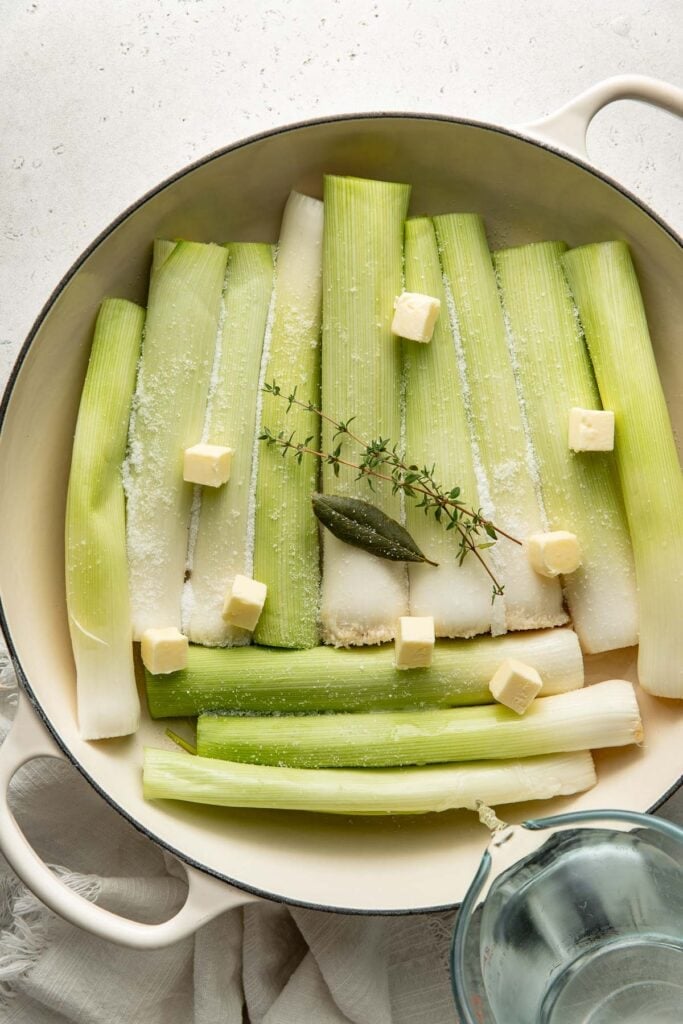 Raw leeks arranged in a white baking dish with cubes of butter, a bay leaf, and a thyme sprig on top. Coarse salt is sprinkled over the leeks, and a glass measuring cup is partially visible nearby.