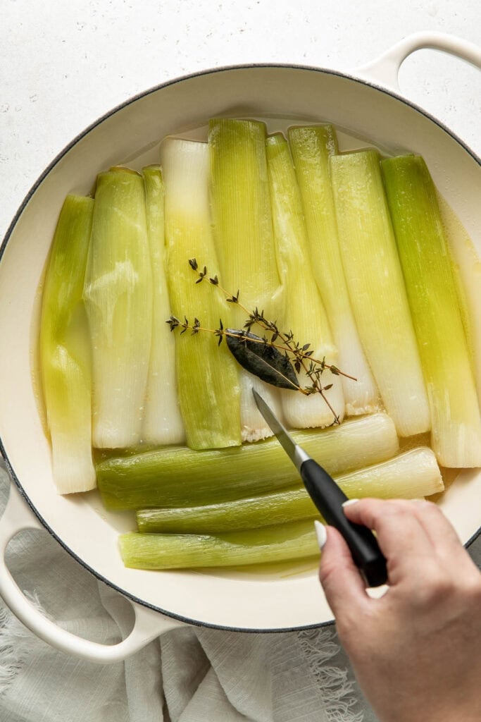 A hand uses tongs to place herbs on top of cooked leeks arranged in a white pan. The leeks are light green and white, and the background is a light, neutral surface.