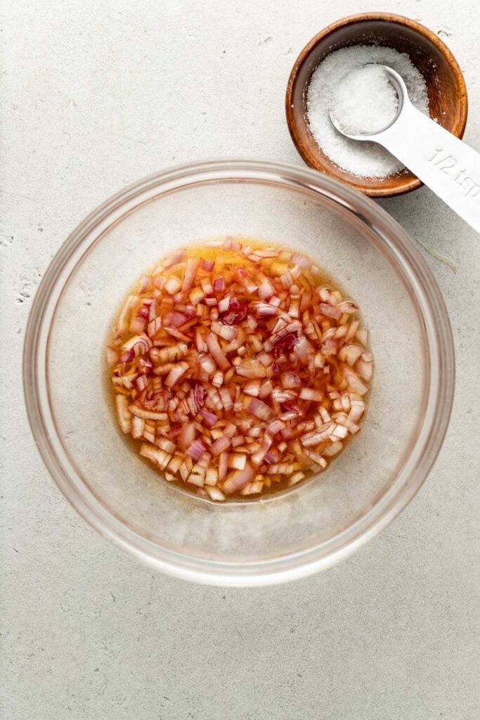 A glass bowl with diced red onions soaking in liquid. Next to it is a small wooden bowl with coarse salt and a white measuring spoon labeled 1/2 tsp. on a light, textured surface.