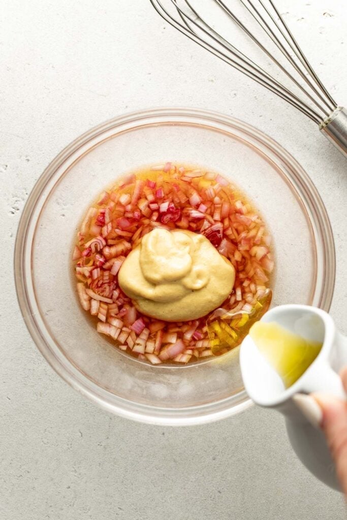 A glass bowl with chopped red onions, mustard, and honey, as olive oil is poured in. A whisk rests nearby on a light surface.