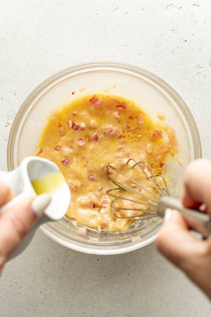 A hand pours oil from a small pitcher into a bowl of whisked eggs and chopped red onions; another hand holds a whisk in the bowl.