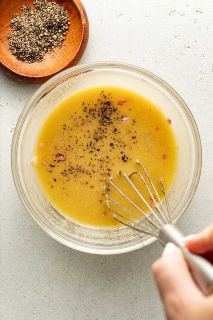 A hand whisks a yellow vinaigrette with chopped onions in a glass bowl. Ground black pepper is sprinkled on top. A small wooden dish with more black pepper sits nearby on a light countertop.